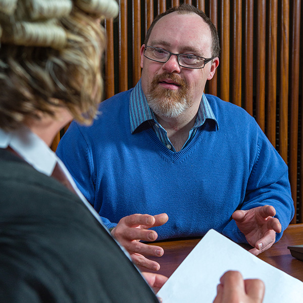 An image of a man wearing blue being cross examined by a lawyer. 